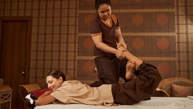Therapist performing a Thai-style assisted stretch on a relaxed woman lying face down on a massage table in a warmly lit room