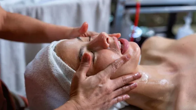 Woman lying back with a towel wrapped around her head receiving a facial massage, hands applying oil to her cheeks and neck.